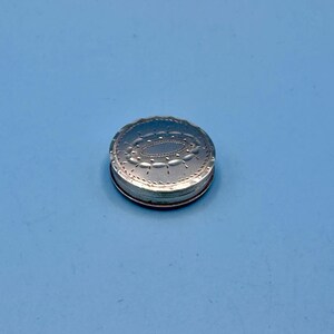 May include: A small, round, silver-coloured container with a hinged lid. The lid features an oval, dotted design surrounded by a decorative border. The container is set against a solid blue background.