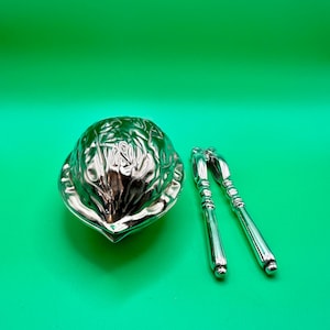 Op de afbeelding: Een glanzende zilverkleurige metalen notenkraker naast een bijpassende zilverkleurige metalen houder in de vorm van een getextureerde walnootschaal. Beide voorwerpen hebben een gepolijst, reflecterend oppervlak met gedetailleerde afwerking.