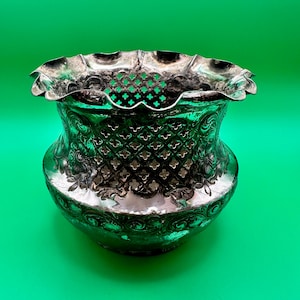 May include: A silver-toned metal bowl featuring a wide, ruffled rim and a decorative lattice-cut pattern around the upper section. The body of the bowl is adorned with embossed floral and scrollwork details, creating a textured, reflective surface.