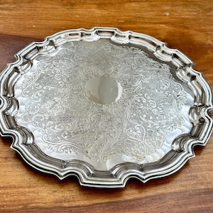 May include: A round silver-toned serving tray featuring an intricate engraved floral pattern across its surface. The tray has a scalloped, raised edge with a polished finish and a smooth, circular blank centre area for potential engraving.