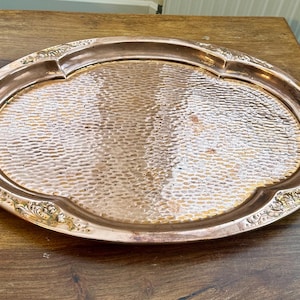 May include: An oval copper serving tray featuring a central hammered texture and a smooth, raised rim with embossed floral detailing. The tray includes two curved brass handles attached to the sides, providing a metallic contrast to the copper.