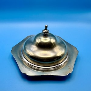 May include: A silver butter dish with a domed lid and a hexagonal base. The lid features a small knob. The dish has a reflective surface, and the base edges are scalloped. The background is a solid blue.