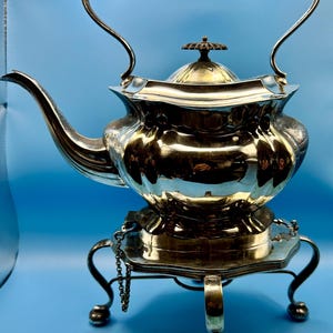 Può includere: Una teiera color argento con manico nero e supporto abbinato. La teiera ha un beccuccio curvo e un coperchio decorativo. Il supporto ha gambe curve e un dettaglio a catena. Lo sfondo è blu.