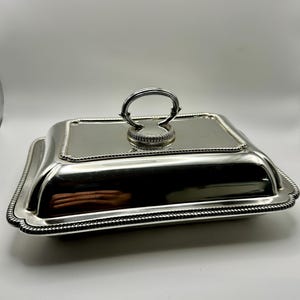 May include: A silver-coloured, rectangular serving dish with a lid. The lid has a rounded handle and a beaded edge. The dish has a beaded edge and a slightly curved shape. The design is classic and elegant.