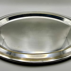 Può includere: Un vassoio ovale argentato con un bordo decorativo. Il bordo presenta anelli concentrici e un motivo strutturato. La superficie del vassoio è molto riflettente, suggerendo una finitura lucida. Il vassoio è progettato per servire cibo o esporre oggetti. Diametro circa 30 cm.
