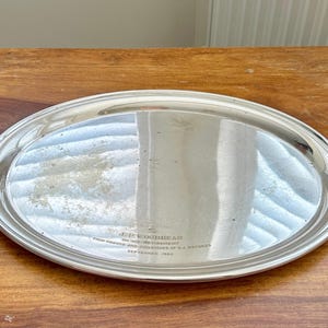 May include: An oval silver-toned serving tray with a polished reflective surface. The tray features an engraved inscription that reads J.P. WOODHEAD ON HIS RETIREMENT FROM FRIENDS AND COLLEAGUES OF B.A. RECORDS SEPTEMBER 1984 centred on the bottom.