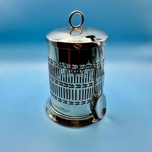 May include: A silver sugar bowl with a lid and spoon. The cylindrical bowl features a decorative lattice pattern. The lid has a ring handle. The spoon is placed on the side. The item is made of silver and is designed for serving sugar.