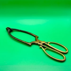 May include: A vintage brass wick trimmer with a scissor-like design, featuring two oval finger loops, a central pivot point, and a long, curved arm that terminates in a small, cup-shaped head for catching trimmed candle wicks.