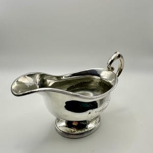 May include: A silver gravy boat with a curved spout and a handle. The boat sits on a round, tiered base. The interior of the boat is reflective, and the exterior is polished.