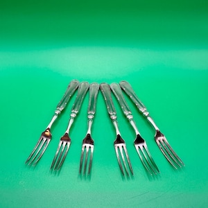 May include: A set of six silver-toned metal forks arranged in a fan shape. Each fork features a slender handle with a decorative embossed line pattern running down the centre and a polished finish on the three-tined head and the handle.