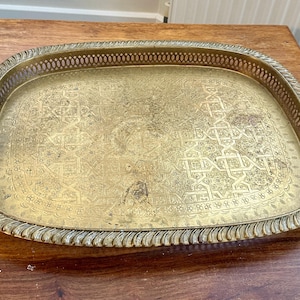 May include: A rectangular brass tray with rounded corners, featuring an intricate engraved geometric pattern on the base and a decorative pierced gallery rim with a twisted rope edge. The metal surface shows a weathered, aged patina throughout.