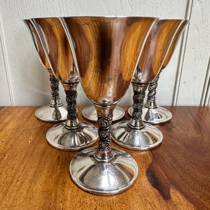 Könnte beinhalten: Sechs silberbeschichtete Kelchgläser mit geriffelter Schale. Die Stiele weisen ein detailliertes Blumenmuster auf, und die Kelchgläser stehen auf runden Sockeln. Das Innere der Kelchgläser hat einen warmen, reflektierenden Ton. Auf Holzoberfläche.