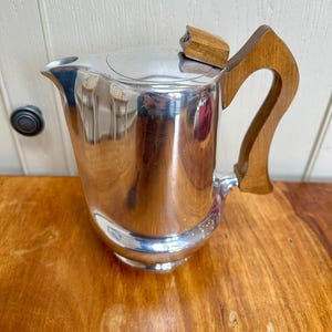 May include: A shiny silver jug with a wooden handle and lid. The jug has a curved spout and a rounded base. The wooden handle is curved and attached to the side. The lid is also made of wood.
