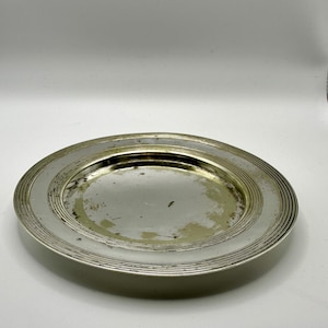 May include: A silver-coloured plate with a circular design. The plate has a wide rim with concentric grooves and a slightly concave centre. The surface shows signs of wear, with some discolouration.
