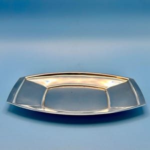 Könnte beinhalten: Ein rechteckiges Tablett aus silberfarbenem Metall mit einer glatten, reflektierenden Oberfläche und leicht erhöhten, abgewinkelten Kanten. Das Tablett verfügt über einen breiten, flachen Rand, der zur Mitte hin abfällt und eine flache Schale bildet.