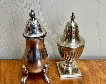 May include: Two antique silver salt and pepper shakers. The salt shaker is a bulbous shape with a lid and three feet. The pepper shaker is urn-shaped with a square base and a perforated lid. Both shakers are highly polished and reflect the light.