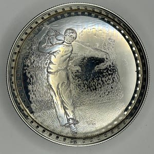 May include: A silver-colored, round tray with a detailed engraving of a golfer mid-swing. The tray has a decorative border with a repeating pattern. The golfer is depicted on a golf course, with trees and foliage in the background.