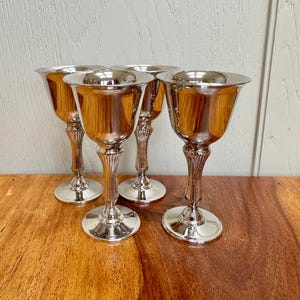 Könnte beinhalten: Vier silberne Kelchgläser mit reflektierender Oberfläche stehen auf einem Holztisch. Die Kelchgläser haben ein klassisches Design mit einer breiten Schale, einem schlanken, geriffelten Stiel und einem kreisförmigen Fuß. Der Hintergrund zeigt eine hellfarbige Wand.