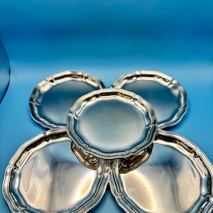 May include: A set of five circular silver-toned plates with a smooth, reflective surface and a scalloped, raised edge. The plates are arranged in a cluster, with one plate positioned centrally and slightly elevated above the four surrounding plates.