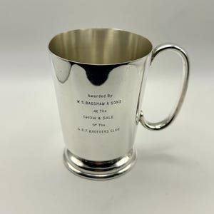 May include: A silver trophy cup with a handle. The cup has text that reads "Awarded By W.S. BAGSHAW & SONS At The SHOW & SALE Of The S.B.F. BREEDERS CLUB". The cup is on a white background.