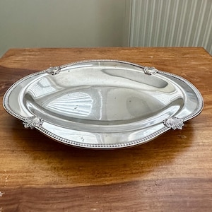 Peut inclure: Un plateau de service ovale en argent avec une surface polie réfléchissante et un bord perlé. Le plateau présente quatre accents décoratifs en forme de coquille positionnés symétriquement le long du bord, créant une bordure détaillée.