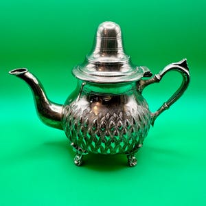Op de afbeelding: Een zilverkleurige metalen theepot met een bolvormig lichaam en een gedetailleerd, in reliëf aangebracht bladerpatroon. De theepot heeft een gebogen tuit, een scharnierend deksel met een puntige knop, een decoratief handvat en drie kleine voetjes.