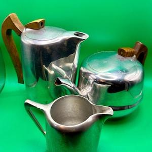 May include: A set of three silver-toned metal vessels including a tall coffee pot with a wooden handle, a shorter rounded teapot with a wooden handle, and a small open creamer pitcher with a metal handle, all featuring smooth metallic surfaces.