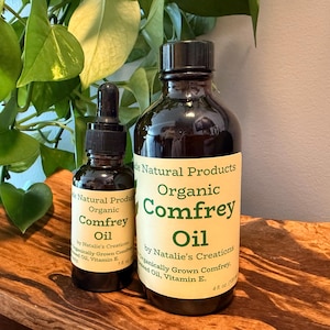 May include: Two dark glass bottles of organic comfrey oil. The larger bottle has a black cap and a label that reads "Organic Comfrey Oil." The smaller bottle has a dropper and a similar label. Both bottles are on a wooden surface.