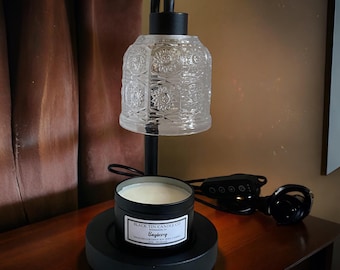 May include: A black candle warmer with a clear, patterned glass shade. A black tin candle labeled "Bayberry" sits on the base. The warmer has a black metal frame and a power cord with a dimmer switch. The candle warmer is on a wooden surface.