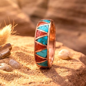 May include: A copper-colored ring with a geometric design. The ring features alternating turquoise and red triangular inlays. The ring is set against a sandy background with small stones and a piece of dried plant.
