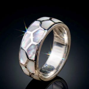 May include: A silver-toned ring featuring a band inlaid with iridescent white mother-of-pearl segments arranged in a geometric honeycomb pattern. The polished metal edges frame the shimmering shell pieces, creating a reflective surface with subtle rainbow hues.