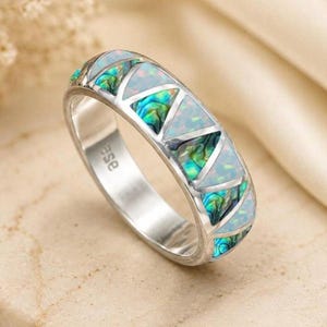 Peut inclure: Une bague argentée dotée d'un anneau avec des incrustations triangulaires alternées d'opale blanche irisée et de coquille d'ormeau aux reflets verts et bleus. L'intérieur de l'anneau porte l'inscription gravée "ase" en minuscules.