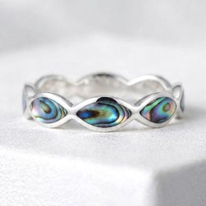 May include: A silver band ring featuring a series of marquise-shaped inlays of iridescent abalone shell. The shell displays swirling patterns of blue, green, and purple hues set within the polished metal frame of the circular jewelry piece.