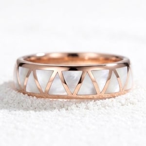 Può includere: Anello in oro rosa con design geometrico. L'anello presenta una fascia in oro rosa con ritagli triangolari riempiti di madreperla bianca. L'anello è posto su uno sfondo bianco strutturato.