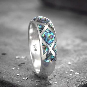 Könnte beinhalten: Ein Silberring mit einem Band, das ein geometrisches Muster aus dreieckigen und rautenförmigen Einlagen aus irisierendem Abalone-Muschelmaterial aufweist. Die Innenseite des Rings ist mit der Zahl 925 für Sterlingsilber gestempelt.