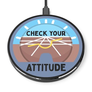 Puede incluir: Un cargador inalámbrico negro con un gráfico azul y marrón que dice "CHECK YOUR ATTITUDE". El diseño se asemeja al indicador de actitud de un avión, con una línea de horizonte amarilla y marcas blancas. El cargador tiene un cable negro.