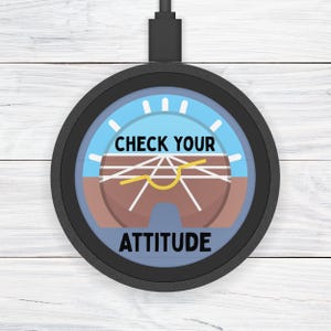 Puede incluir: Un cargador inalámbrico negro con un diseño de indicador de actitud de avión. El indicador muestra un cielo azul, suelo marrón y el texto "CHECK YOUR ATTITUDE".