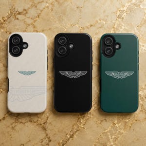 May include: Three smartphone cases in white, black, and dark green, each featuring a circular camera cutout and the Aston Martin winged logo. The white case has a textured surface, while the black and green cases have a smooth, matte finish.