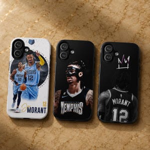 May include: Three smartphone cases featuring basketball player Ja Morant. The left case is white with a blue Grizzlies jersey graphic and text. The middle case is black with a masked player portrait. The right case is black with a jersey back graphic.