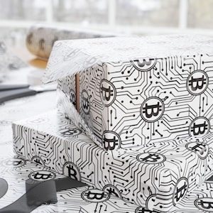 May include: Two gift boxes wrapped in white paper with a black circuit board and Bitcoin symbol pattern. A roll of matching wrapping paper, scissors, and a gray ribbon are also visible, suggesting a theme of cryptocurrency or technology.