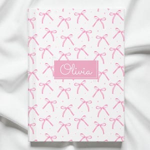 May include: A white notebook with a repeating pattern of pink bows and small diamonds. The name "Olivia" is displayed in cursive within a pink rectangular box on the cover. The notebook is lying on a white fabric surface.