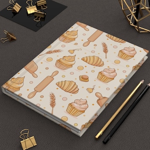 May include: A cream-coloured journal with a repeating pattern of baked goods. The design includes cupcakes, croissants, rolling pins, and wheat stalks in shades of tan and peach. Gold and black pencils and gold binder clips are also visible.