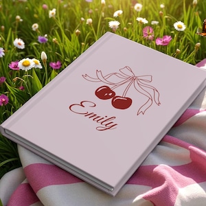 Może przedstawiać: Jasnoróżowy notatnik z motywem wiśni i imieniem "Emily" w kursywie. Wiśnie są w głębokim czerwonym kolorze, a wstążka w jaśniejszym odcieniu czerwieni. Notatnik leży na różowo-białym pasiastym kocu.