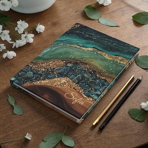May include: A hardcover journal with a unique design featuring shades of teal, gold, and brown. The cover has a marbled effect with the name "Sofia" in elegant script. Two pencils, one gold and one black, are placed beside the journal.