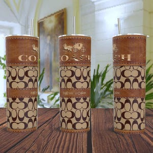 Puede incluir: Tres vasos marrones y beige con un patrón repetido y el texto "COACH NEW YORK". Cada vaso tiene una banda marrón similar al cuero en la parte superior e inferior con un logotipo y una pajita plateada.