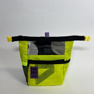 Könnte beinhalten: Eine neongelbe und schwarze Tasche mit Reißverschluss oben und einer kleinen Vordertasche. Die Tasche hat einen schwarzen Riemen mit gelben Schnallen an jedem Ende. Der Text "BUG BAG" befindet sich auf einem lila Aufnäher auf der Vorderseite.