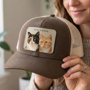 May include: A brown trucker cap with a beige mesh back. The front patch displays a portrait of two cats, a calico and an orange tabby, with the text "LUNA & MOOSE".