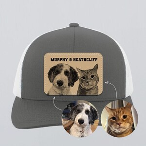 May include: A gray and white trucker hat with a white mesh back. The front features a leather patch with a sepia-toned portrait of a dog and a cat, and the text "MURPHY & HEATHCLIFF". Below are two circular pet portraits.