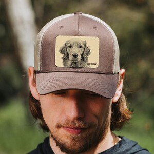 May include: A brown trucker hat with a beige mesh back and a rectangular patch. The patch features a detailed black and white illustration of a Golden Retriever dog with the name "BUDDY" below.