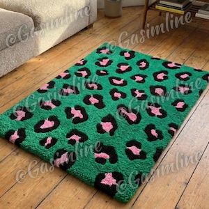 Puede incluir: Una alfombra rectangular verde con un diseño de estampado de leopardo. La alfombra presenta manchas de leopardo negras y rosas sobre un fondo verde. La alfombra está colocada sobre un suelo de madera.